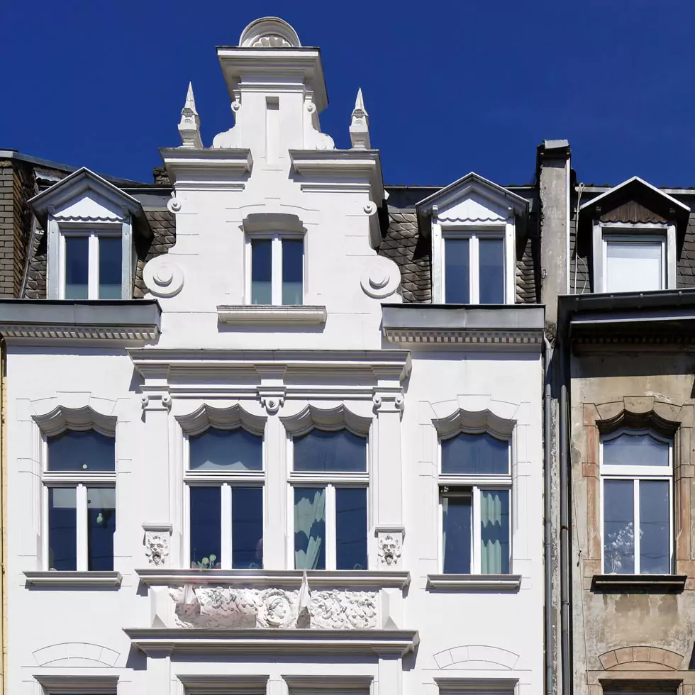 Fassade eines sanierten Altbaus mit weißer Stuckverzierung, mehreren Fenstern und Dachgauben unter blauem Himmel.