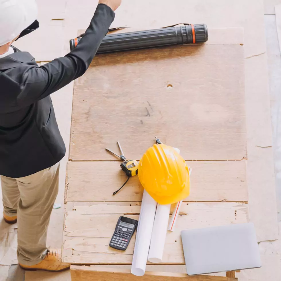 Person mit weißem Helm und schwarzer Jacke steht an einem Holztisch mit gelbem Bauhelm, Bauplänen, Taschenrechner und Laptop.
