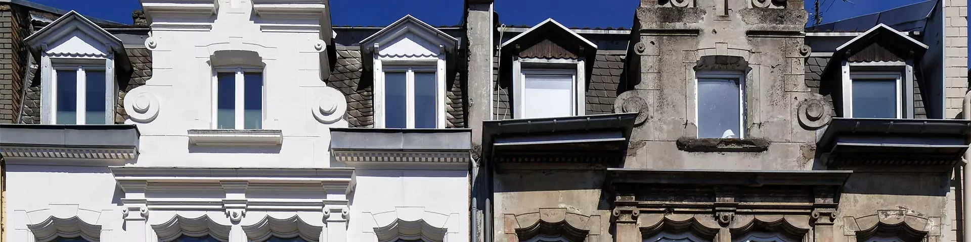 Fassade eines historischen Gebäudes mit weißen und grauen Teilen, Fenstern und Dachgauben unter blauem Himmel.