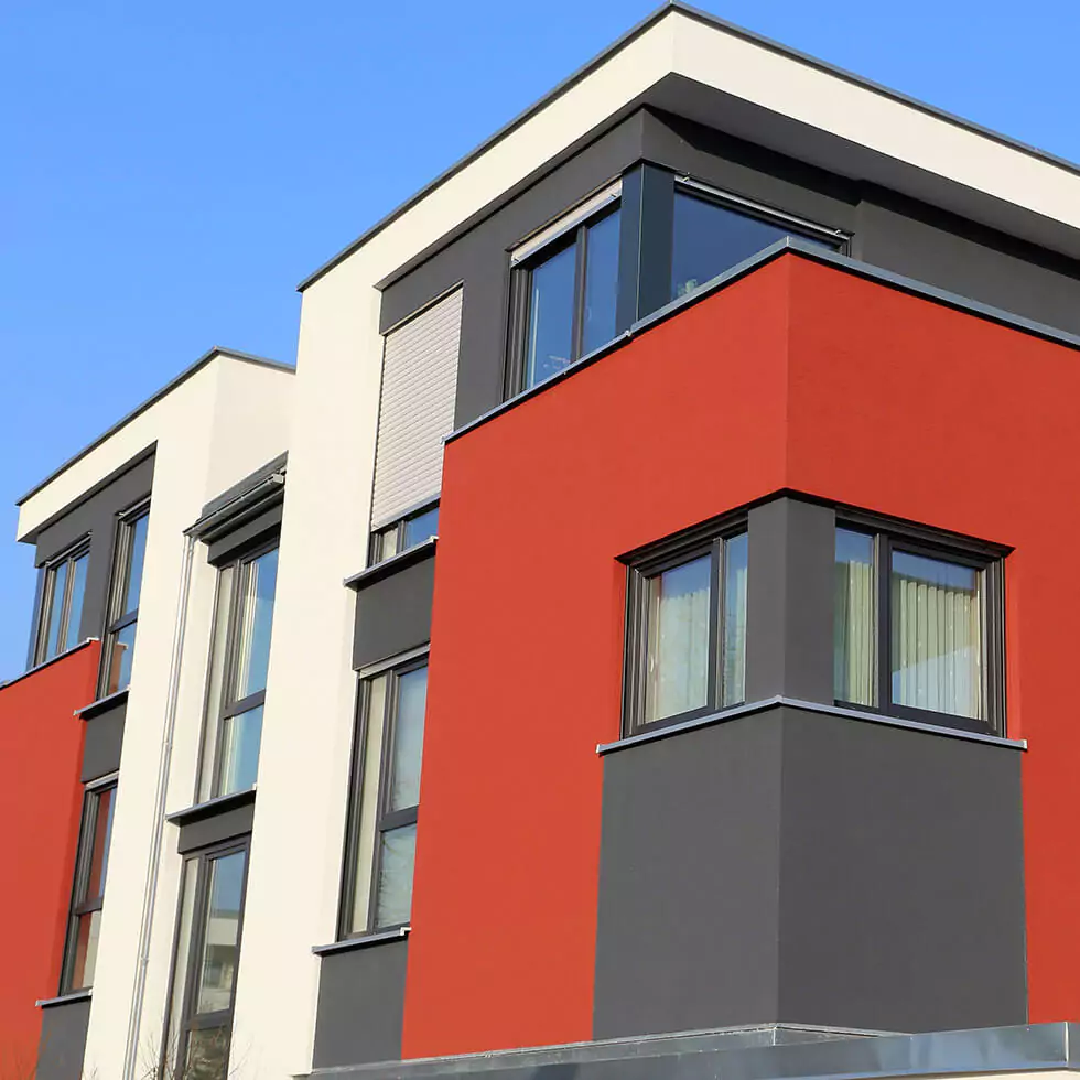 Moderne Gebäudeecke mit roten, grauen und weißen Fassadenflächen und mehreren Fenstern unter blauem Himmel.