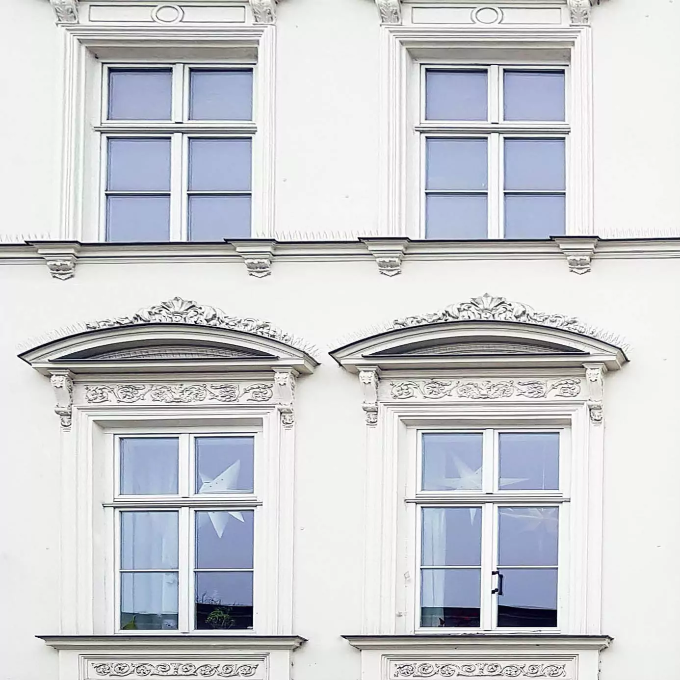 Fassadenansicht eines weißen Gebäudes mit drei Fenstern und dekorativen Stuckelementen über den Fenstern.