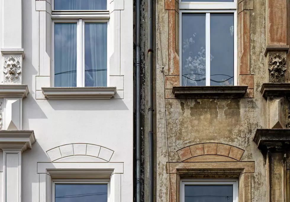 Zwei nebeneinanderliegende Hausfassaden, links frisch renoviert mit weißen Fenstern, rechts alt und verwittert mit dunklen Fenstern.