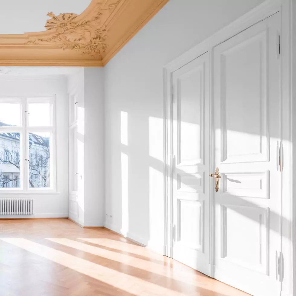 Heller Raum mit weiß gestrichenen Wänden und Türen, hellbraunem Parkettboden und verziertem, goldfarbenem Stuck an der Decke.