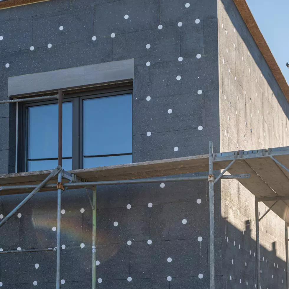 Fassade mit grauen Wärmedämmplatten, die mit gelben Dübeln befestigt sind, neben einem Baum und einem Zaun.