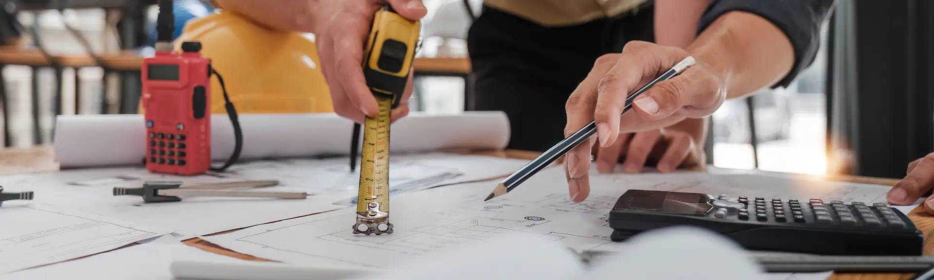 Hände von zwei Personen, eine hält ein Maßband über Bauplänen, die andere zeigt mit einem Stift darauf, daneben Taschenrechner und Bauhelm.