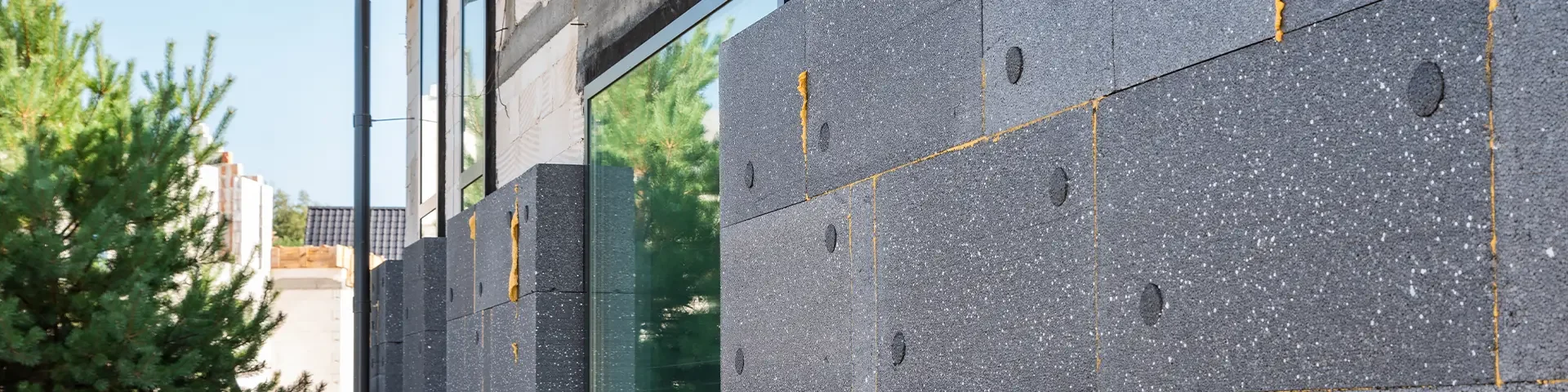 Fassade mit grauen Wärmedämmplatten, die mit gelben Dübeln befestigt sind, neben einem Baum und einem Zaun.