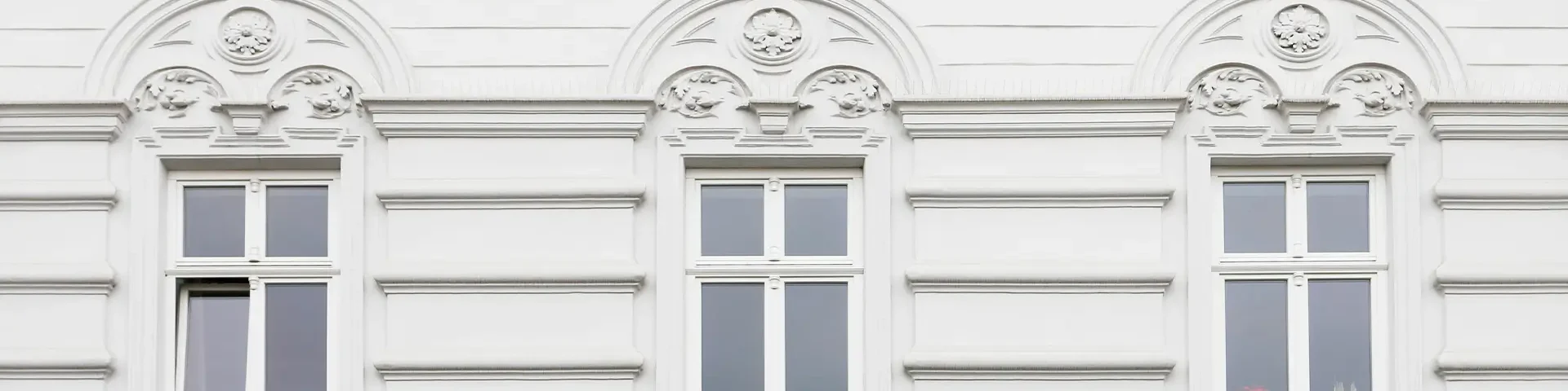 Fassadenansicht eines weißen Gebäudes mit drei Fenstern und dekorativen Stuckelementen über den Fenstern.
