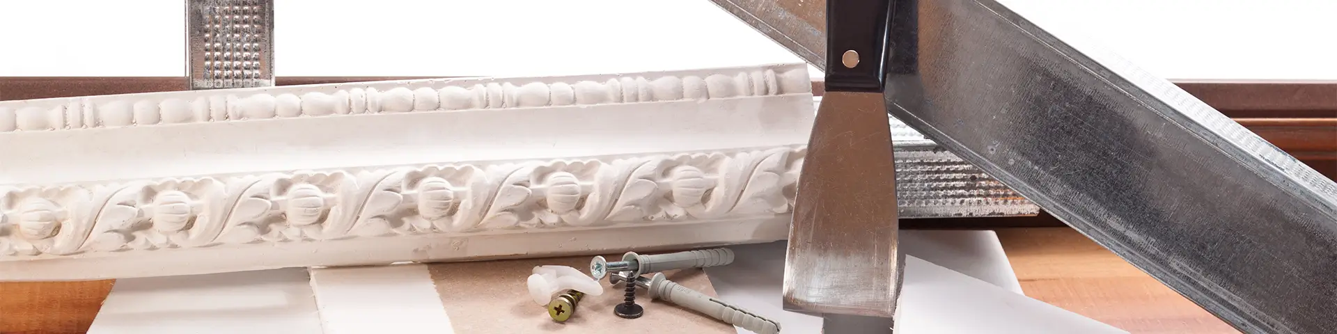 Werkzeug mit schwarzem Griff und Metallklinge liegt auf weißer Stuckleiste mit floralem Muster auf Holzboden.